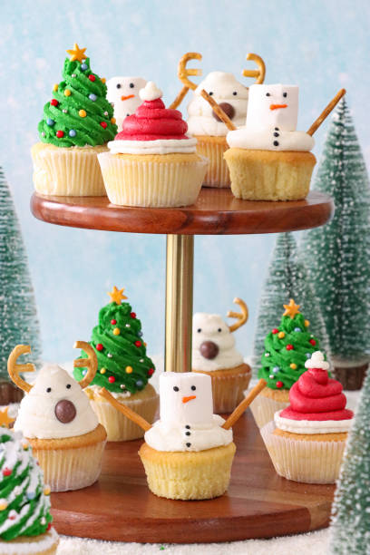 Close-up image of batch of homemade, Christmas design cupcakes, Christmas trees, snowmen, Santa hats and reindeers decorated cakes on cake stand, icing sugar snow, pale blue background, focus on foreground