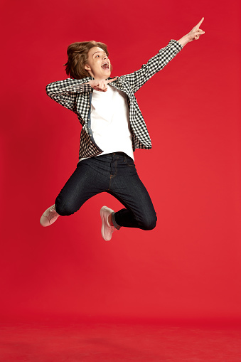 Dance, sing song, favourite melody. Exciting yound boy, student wearing checkered shirt jumping and dancing with hands over red background. Concept of music, hobby, human emotions, mood, ad