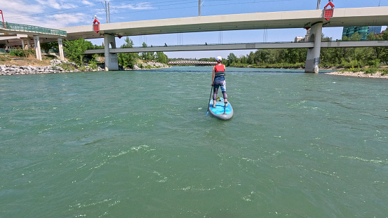 Woman SUP boarder paddles down river towards bridges in urban environment