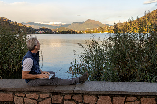 Senior man sits on wall above lake and works on laptop