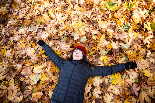 A Caucasian woman in her late 50's enjoys exploring a forest area adjacent to the Puget Sound in Washington state.  The fall leaves add a vibrant yellow, orange, and brown color pop to the scene.  She lays on her back in piles of fallen leaves.