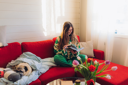 Smiling female in green clothing sitting on comfortable sofa with her cute pug who is sleeping, contemplating winter day indoors reading book with bouquet of tulip flowers in vase and mountain window view