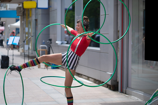 A street performer is spinning two hoops on one arm and two hoops on her other arm, and a hoop of each leg while standing on one leg on a hot day in the city