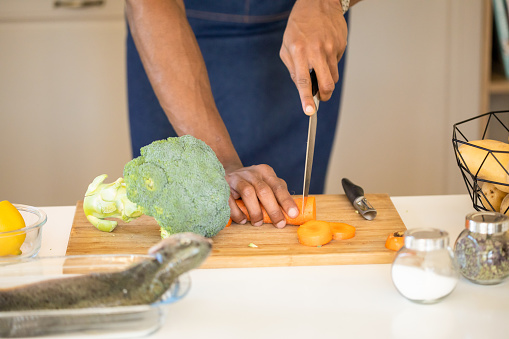 Close up of a kitchen table, with broccoli on a cutting board next to a carrot being cut with a sharp knife by a pair of hands, a raw fish in a dish, and salt and pepper in little jars a fresh lemon and some raw potatoes