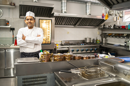 A male Asian chef wearing bright white's standing by the stainless steel counter with his arms crossed, surrounded by kitchen equipment in a commercial  kitchen