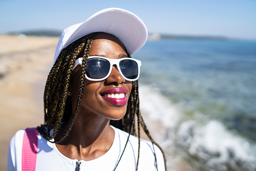 Headshot of a beautiful African-American woman is smiling into the distance, she is wearing a baseball cap and sunglasses as she stands beside the shoreline on a bright sunny day
