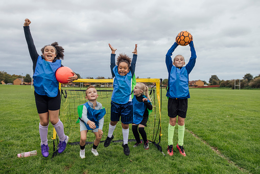 Front view of a small group of boys and girls wearing sports clothing, football boots and a sports bib on a football pitch in the North East of England. They are at football training where they are doing different football training drills. They are jumping up and celebrating with their arms raised in front of a football goal.\n\nVideos are available for this scenario.