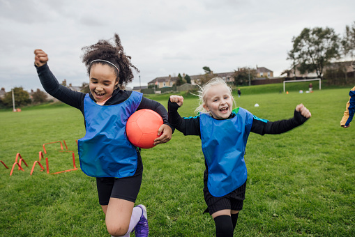 Front view of two young girls wearing sports clothing, football boots and a sports bib on a football pitch in the North East of England. They are at football training where they are doing different football training drills. They are running with their arms raised while they celebrate.\n\nVideos are available for this scenario.