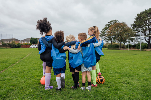Rear view of a small group of boys and girls wearing sports clothing, football boots and a sports bib on a football pitch in the North East of England. They are at football training where they are doing different football training drills. They are standing in a line and they have their arms around each other, they are having a pep talk to motivate them to win before their five-a-side game of football.\n\nVideos are available for this scenario.