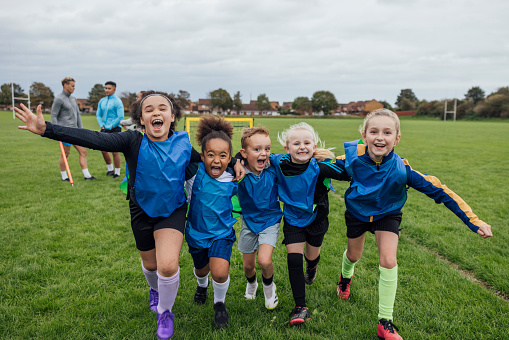 Front view of a small group of boys and girls wearing sports clothing, football boots and a sports bib on a football pitch in the North East of England. They are at football training where they are doing different football training drills. They are running and celebrating with their arms around each other in front of a football goal while smiling and looking at the camera.\n\nVideos are available for this scenario.