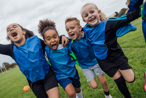 Front view of a small group of boys and girls wearing sports clothing, football boots and a sports bib on a football pitch in the North East of England. They are at football training where they are doing different football training drills. They are running and celebrating with their arms around each other while smiling and looking at the camera.\n\nVideos are available for this scenario.