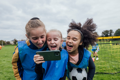 Front view of a small group of girls wearing sports clothing, football boots and sports bibs on a football pitch in the North East of England, they are at football training and they are taking a selfie on a smartphone, they are pulling funny faces.

Videos are available for this scenario.