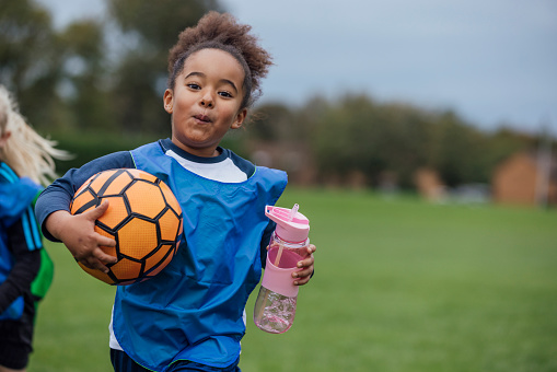 Front view waist up of a young girl running on the grass and holding a water bottle and football while at football training for a children's team in the North East of England. She is wearing sports clothing, football boots and a sports bib.