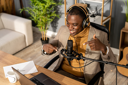 Looking down to an African-American radio presentor gesturing with his hands as he talks into a large microphone, wearing headphones over his ears and sitting at a desk with a laptop open in front of him in a modern studio