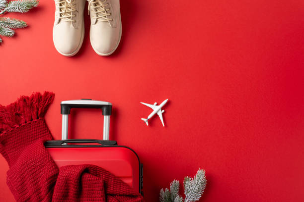 istock Winter Travel Dreams: top-down view featuring white female winter boots, suitcase, a small airplane model, knitted scarf, frosty fir twigs against red background, leaving space for text or advertising 1804198136