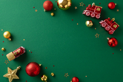 Festive preparations underway. Top view of charming red and gold ornaments, train and house-shaped tree decorations, confetti on a green surface. Ideal for holiday greetings