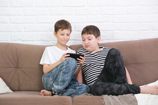 Close up of two cute boys using smartphone, looking at screen, childs holding phone in hands, sitting on couch at home , playing mobile device game, watching cartoons online.