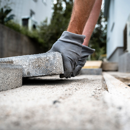 Low angle closeup view of male hands in protective work gloves laying cement tiles to pave a surface around a house.