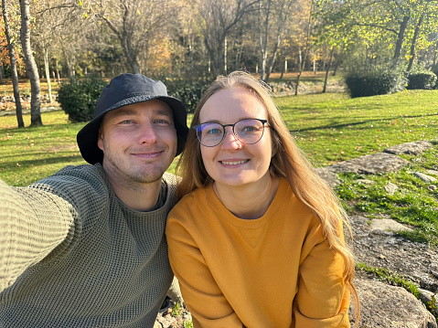 Man in hat and woman in glasses grimacing while taking selfie in park.