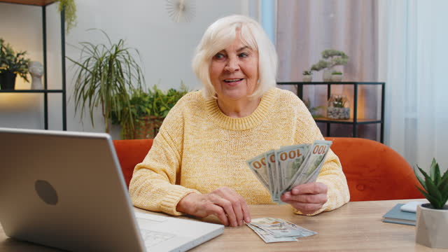 Happy senior woman counting money cash and use laptop pc calculate domestic income earnings at home
