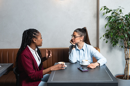 Two young business woman colleagues taking a break in nearby cafeteria drinking coffee talking about private life, to get know each other better. Staff members company employees sitting in the cafe