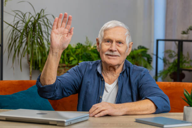 istock Senior man smiling friendly at camera, waving hands gesturing hello, hi, greeting at home on couch 1804261197