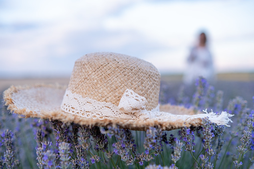 In the foreground a straw hat on a tuft of lavender. In the background, a young woman in a white dress and a romantic sky. The focus is on the hat.