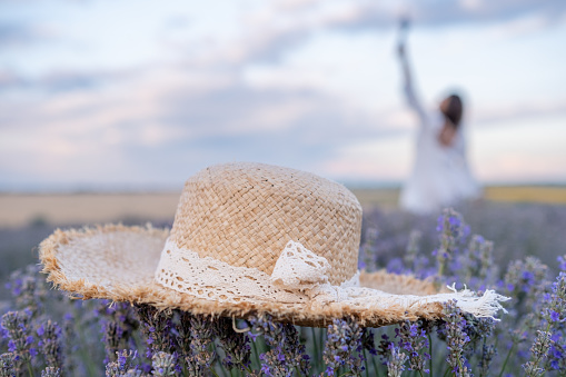 In the foreground a straw hat on a tuft of lavender. In the background, a young woman in a white dress and a romantic sky. The focus is on the hat.