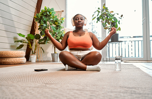 African young lady listening relaxing music in wireless headset while meditating in lotus pose on floor. Peaceful curvy woman keeping eyes closed and holding hands in mudra gesture.