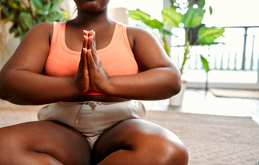 Close up of overweight african woman keeping hands in namaste gesture and sitting on lotus pose while meditating at home, copy space. Concept of yoga, wellbeing and spirituality.