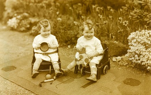 Twin baby girls sitting outside in chairs in a garden playing with toys. Reigate, England 1930.