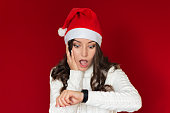 a young pretty woman with curly hair, dressed in a Santa Claus hat  looks at the clock with surprise, clutching her head with her hand and opening her mouth, is late