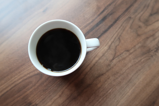 White cup of black coffee on a wooden table surface. Natural light. No people. Morning coffee, soft light. Picture taken from above.
