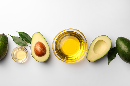 Cooking oil in bowl and fresh avocados on white background, flat lay