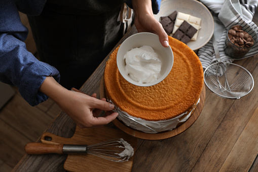 Woman smearing sides of sponge cake with cream at wooden table, closeup