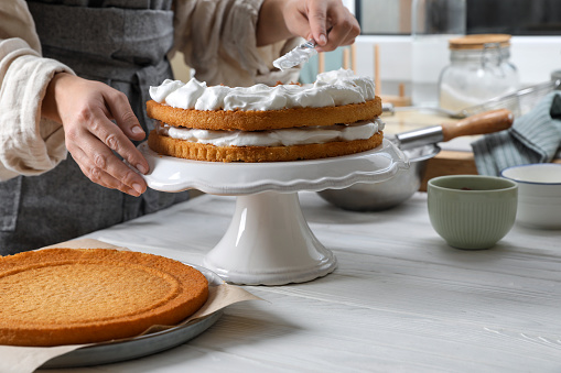 Woman smearing sponge cake with cream at white wooden table in kitchen, closeup