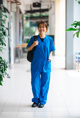 Happy Asian Female Nurse in Scrubs Walking Through Hospital Corridor Carrying a Laptop Bag. Looking at camera.
