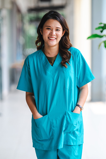 Happy young Asian nurse walks in hospital corrido in scrubs. Looking at camera.