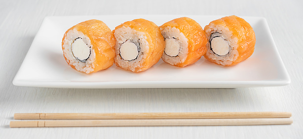 Portion of japanese delicious philadelphia sushi roll made with raw or smoked salmon fish, cream cheese, nori seaweed and boiled rice served on plate with bamboo chopsticks on white wooden table
