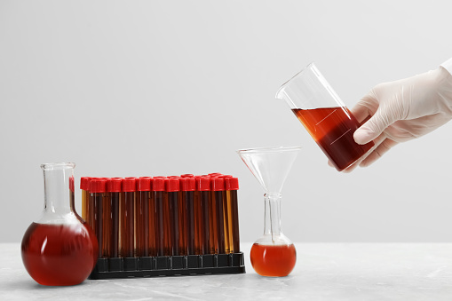 Scientist pouring liquid from beaker into conical flask on white table against light background, closeup