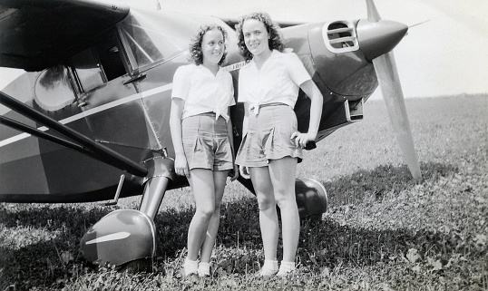 Twin teenage girls standing in a field by a Stinson Voyager airplane. Pennsylvania, USA 1948.