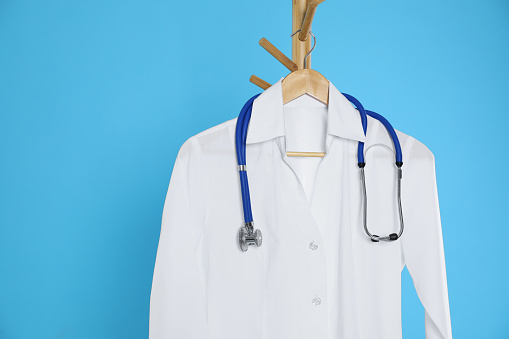 White medical uniform and stethoscope hanging on rack against light blue background. Space for text