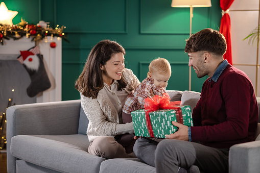 Beautiful parents with their cute little daughter enjoying together at home during Christmastime. Mother is holding a Christmas present