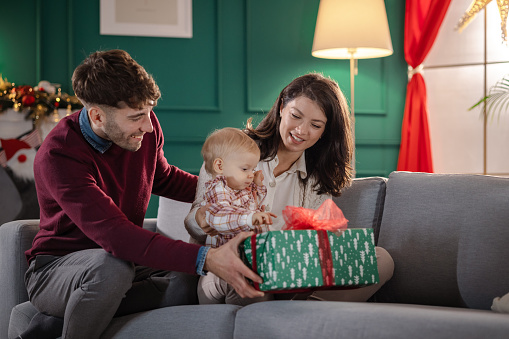 Beautiful parents with their cute little daughter enjoying together at home during Christmastime. Father is holding a Christmas present