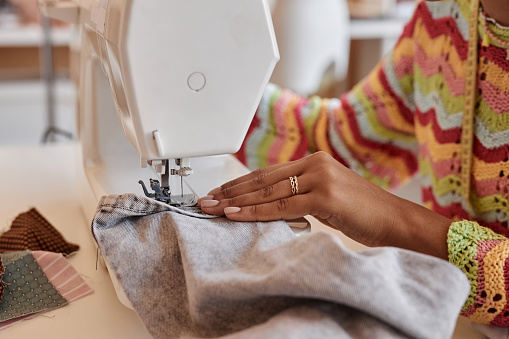 Hand of young African American woman sewing denim clothes while sitting in front of electric machine and creating new fashion items