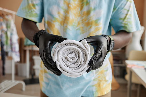 Hands of young African American craftsman in protective gloves holding rolled white t-shirt in front of himself while going to make tie dye