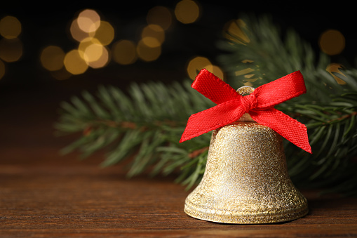 Bell with red bow and fir branches on wooden table against blurred background, closeup. Christmas decor