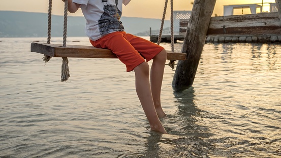 Closeup of boy's feet touching sea waves while swinging on swing at the ocean beach on sunset. Holiday, summer vacation and tourism.