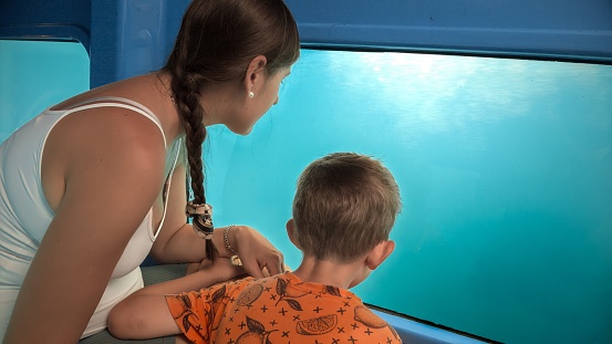 Mother with little son exploring underwater sea world and fishes while traveling on tourist submarine. Holiday, summer vacation and tourism.