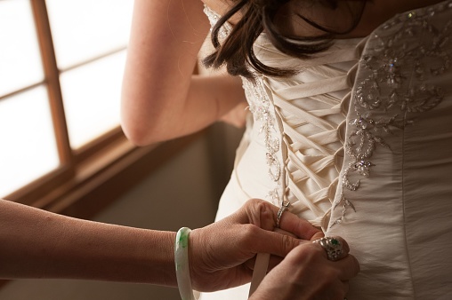 A closeup of a woman's hand tying knots on a bride's wedding dress providing assistance and support during the preparation